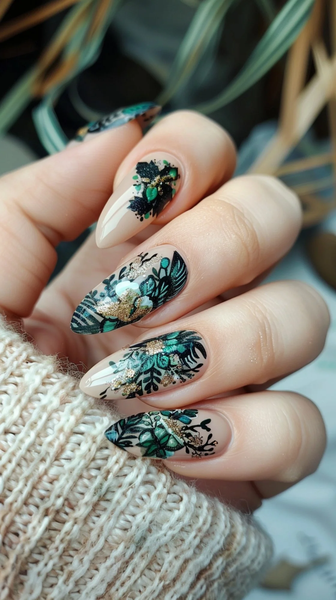 Sophisticated nails with a nude base, adorned with dark teal floral and leaf patterns, accented by luxurious gold flakes and tiny green embellishments, giving a rich, nature-inspired bohemian look.