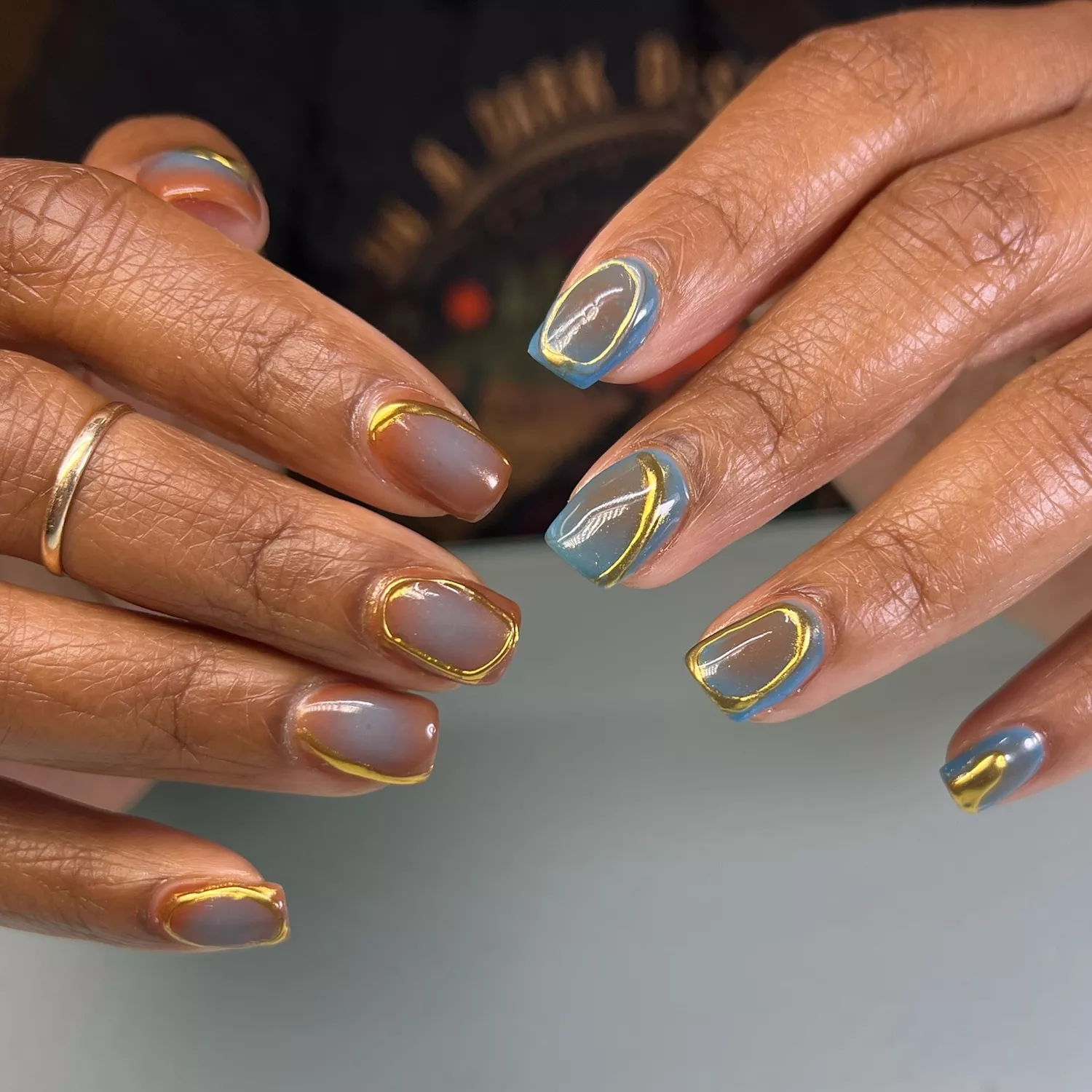 Hand with blue and burnt orange airbrushed nails with raised gold chrome halos on each finger.