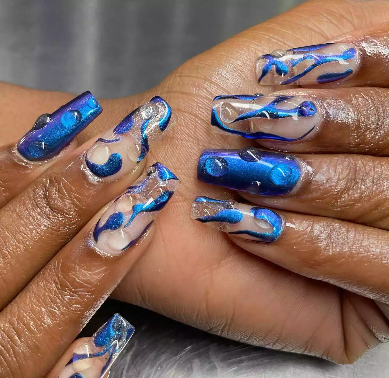 Hand with long square nails with royal blue chrome polish with clear 3D drops.