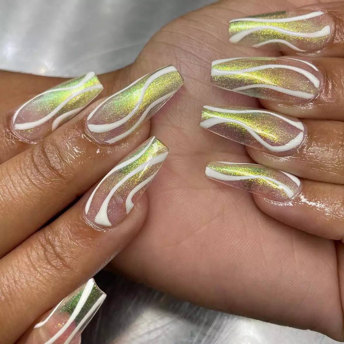 Hand with long square nails with a yellow ombre chrome with white swirls painted throughout the nails .